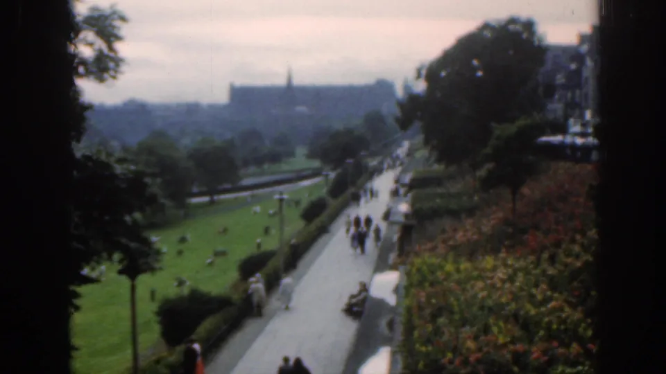 Hazy 1961 Scotland park view from above, showing people on a broad path, green lawns, trees, flower beds, and distant buildings.