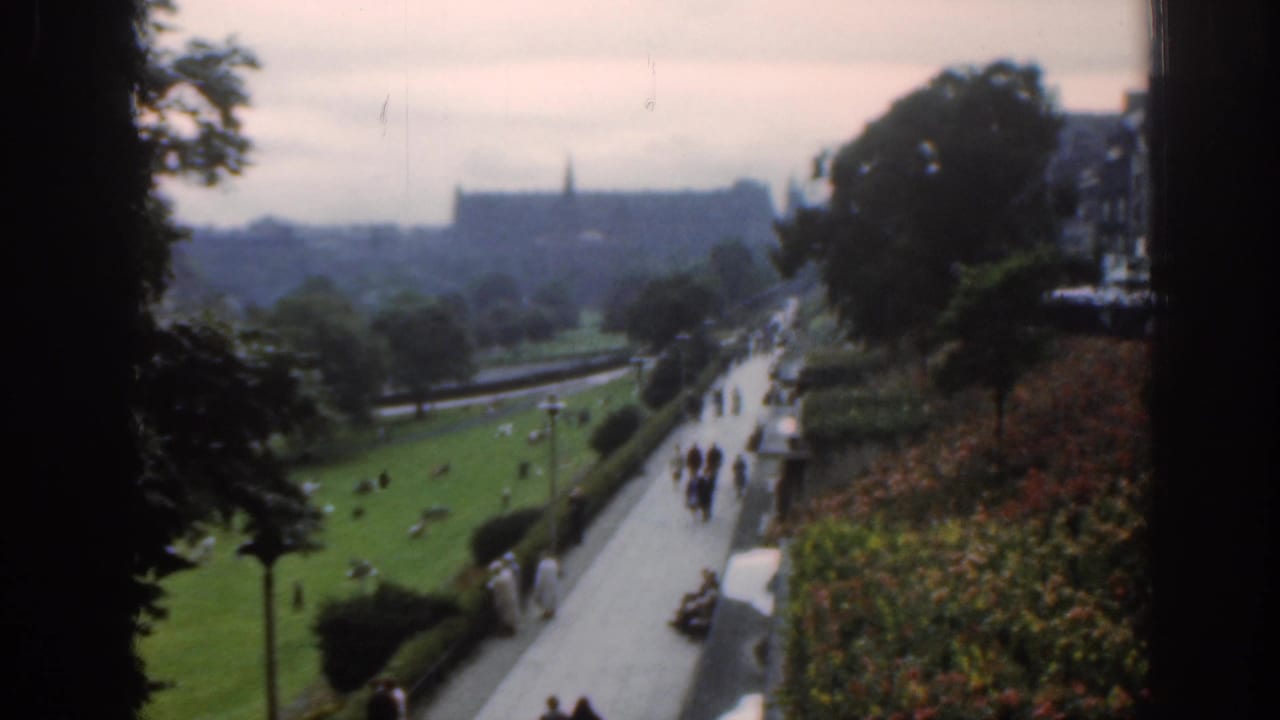 Park path with people strolling beside lawn and flowerbeds, with a distant castle-like building beyond trees.