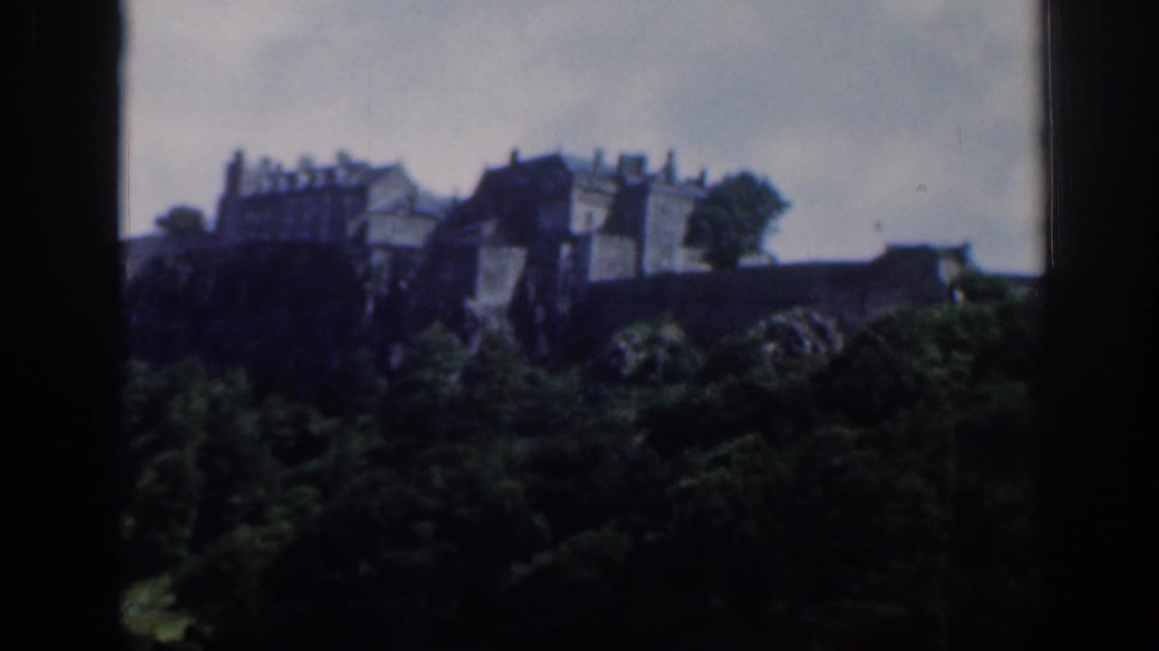 Distant stone buildings on a rocky hill above dense trees, framed by dark film edges.