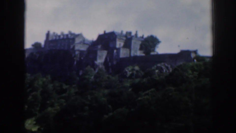 Soft 8mm view of a castle-like structure on a rocky rise above trees in Scotland, with dark foreground framing.
