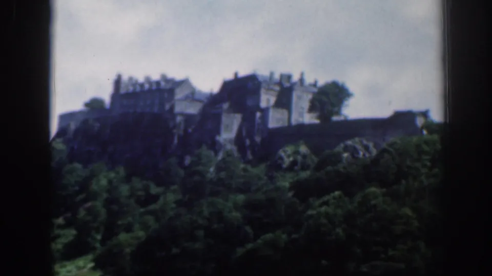 1961 Scotland footage frames a distant historic building complex above dark green trees, with visible 8mm softness and grain.