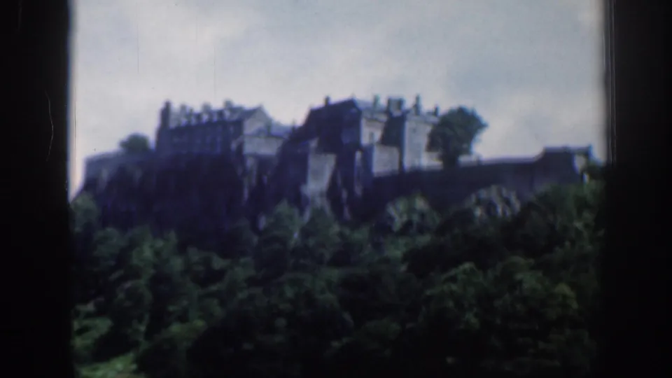 1961 Scotland 8mm frame showing Edinburgh Castle on a rocky rise beyond green trees and fields under a cloudy sky.