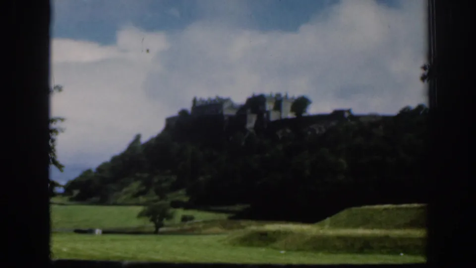 1961 Scotland home-movie frame with Edinburgh Castle seen across parkland, partially silhouetted against a bright cloudy sky.