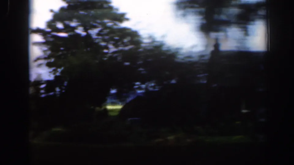 Motion-blurred 8mm roadside frame in Scotland, showing trees, grassy ground, and a dark building or structure partly obscured by shadow.