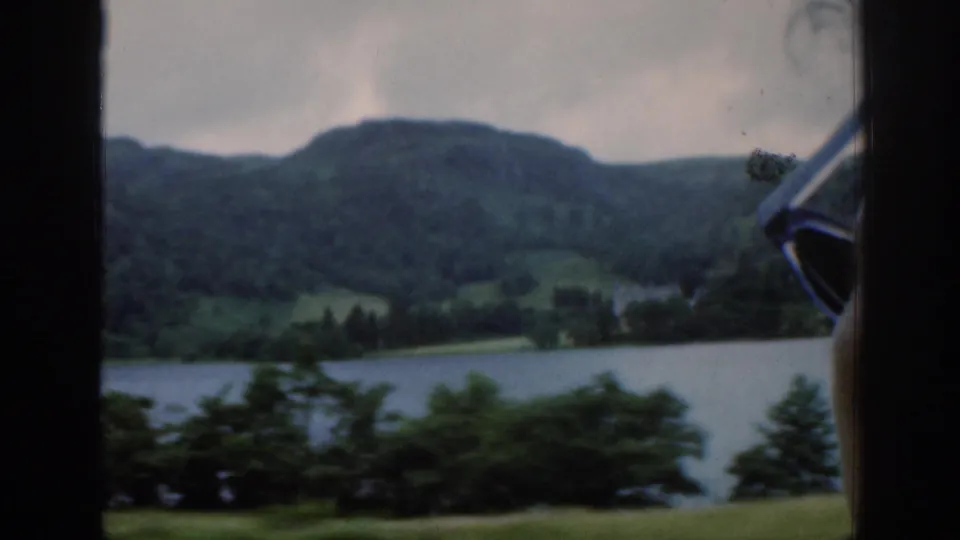 1961 Scotland travel footage frame with a loch, wooded shoreline, and green hills viewed from a moving car.