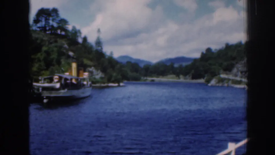 Visitor-shot 1961 Loch Katrine frame looking over blue water toward a steamboat, wooded banks, and low Scottish hills.
