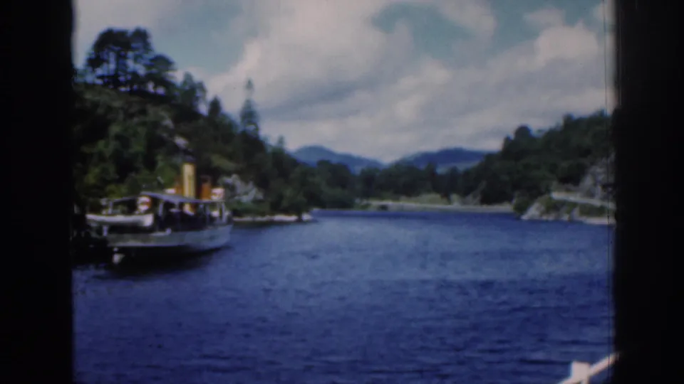 1961 Scotland home-movie frame of a steamboat on Loch Katrine, framed by dark film edges, wooded banks, and distant hills.