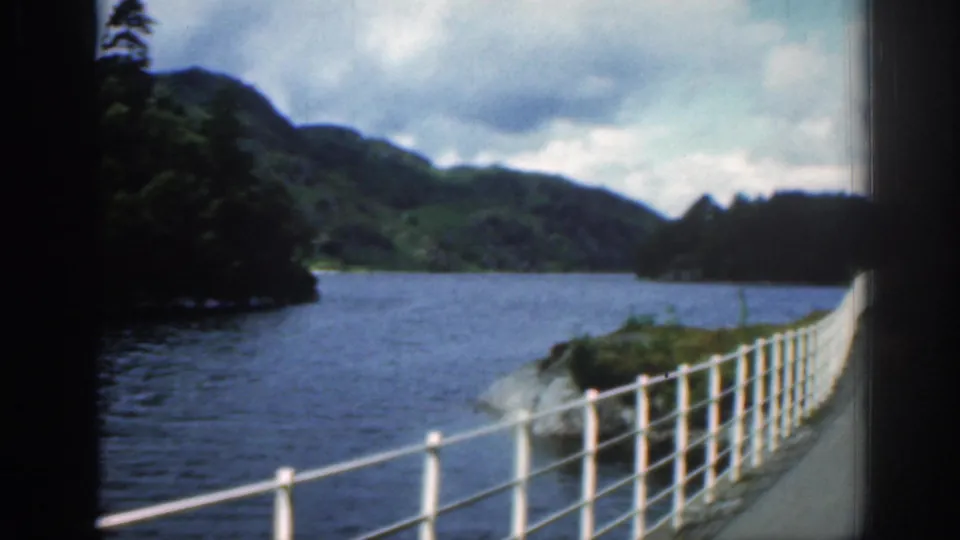 1961 Scotland frame looking along a river or loch edge, with white railings, wooded hills, and cloudy sky in view.