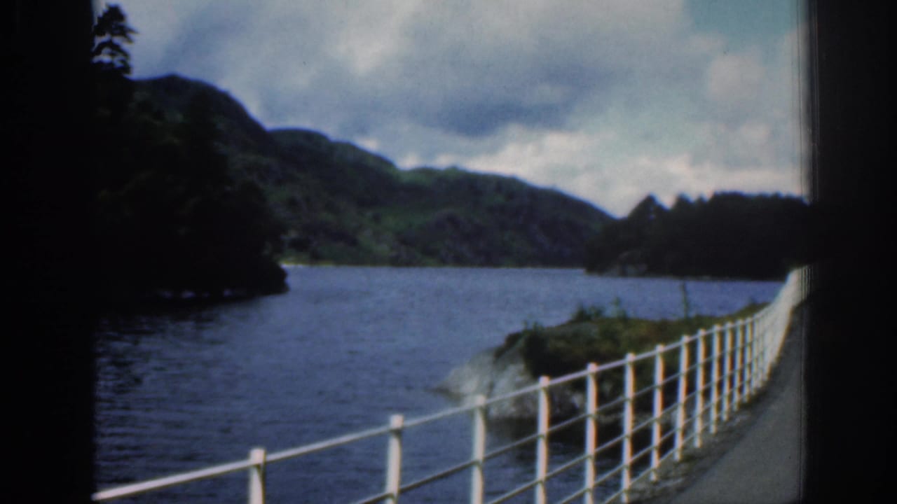 River or loch with green hills, cloudy sky, and white roadside railing.
