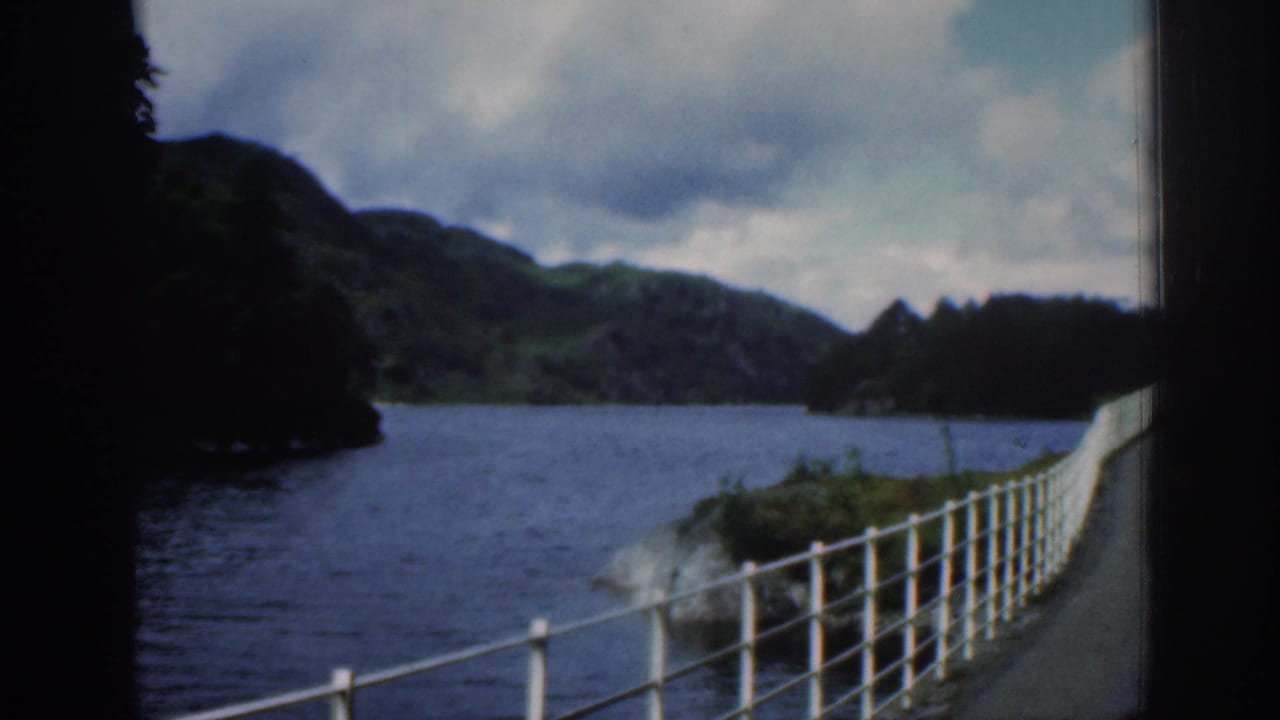 1961 Scotland travel frame with a white metal railing, dark trees, blue water, and hills below a clouded sky.