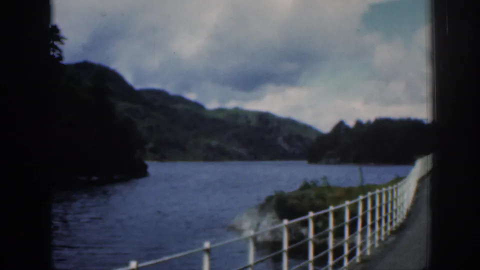 1961 Scotland 8mm frame showing a railing beside a river or loch, with wooded shore, hills, and heavy clouds.