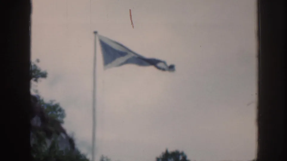 1961 Scotland frame of a blue-and-white Scottish saltire flag fluttering on a pole above trees and rock.