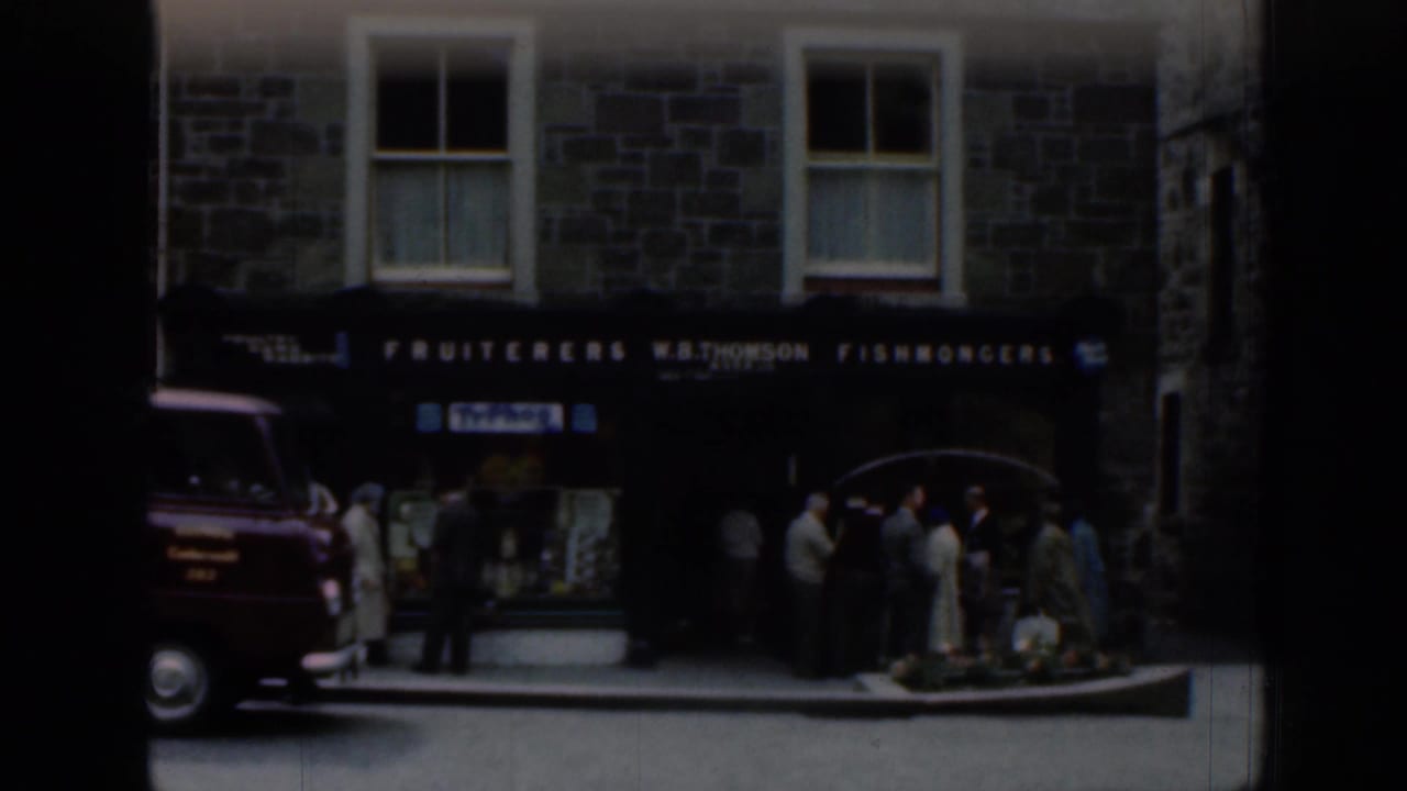 Scottish stone building with W.B. Thomson fishmongers signage, a red delivery van, and townspeople on the sidewalk in 1961 footage.