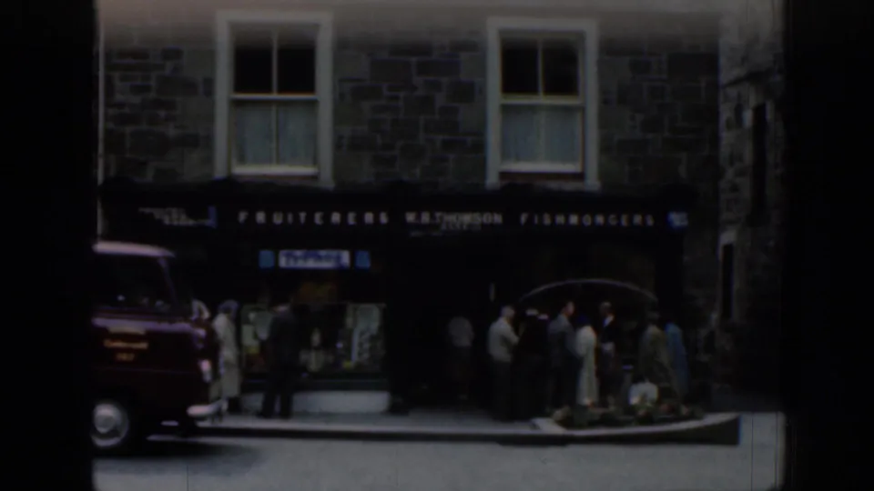 Scottish stone building with W.B. Thomson fishmongers signage, a red delivery van, and townspeople on the sidewalk in 1961 footage.
