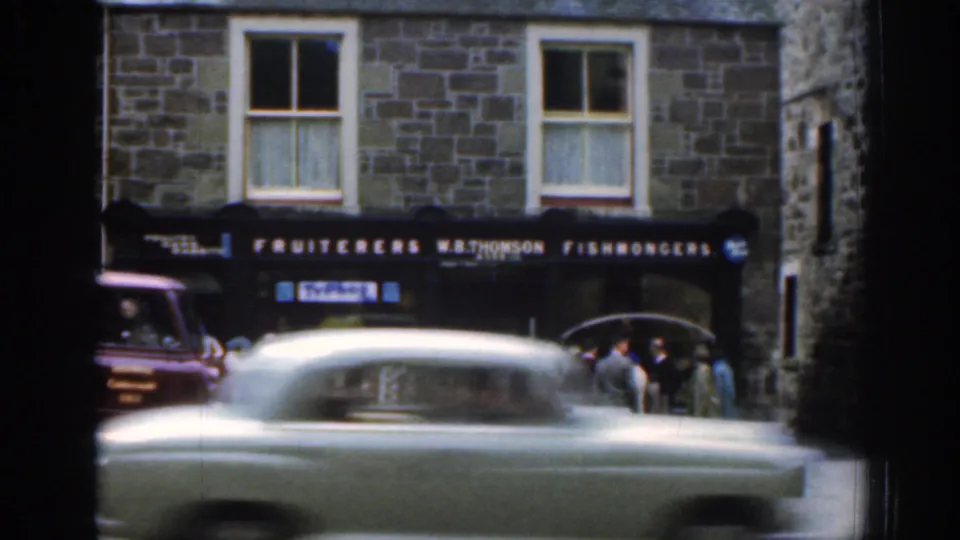 A passing car blurs across the foreground of a Scottish fishmongers shopfront with people and a red van visible behind it.