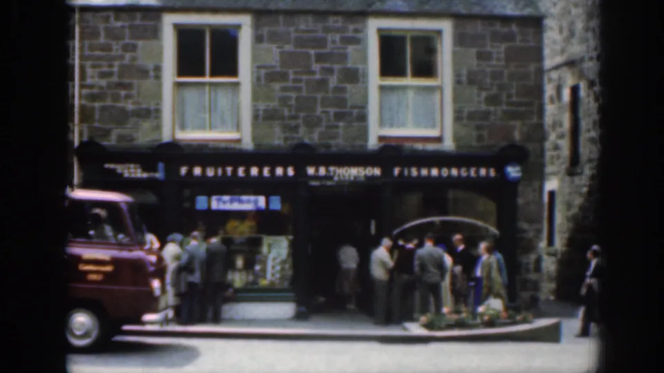 W.B. Thomson fishmongers shop in Scotland with a red van, gathered townspeople, and street goods visible near the curb in 1961 footage.