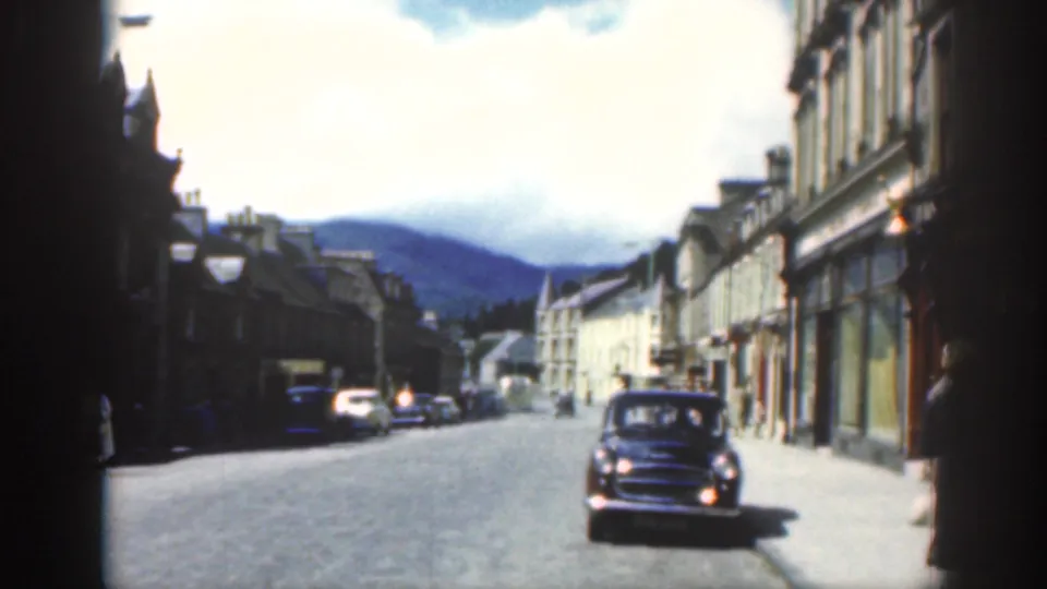 1961 Scotland home-movie street scene with a compact car, parked vehicles, shopfronts, and distant hills.