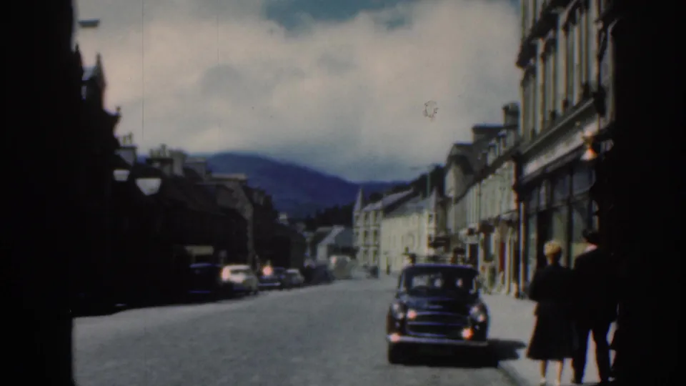 1961 Scotland street view with classic cars, pedestrians at frame edge, shop buildings, and distant hills.