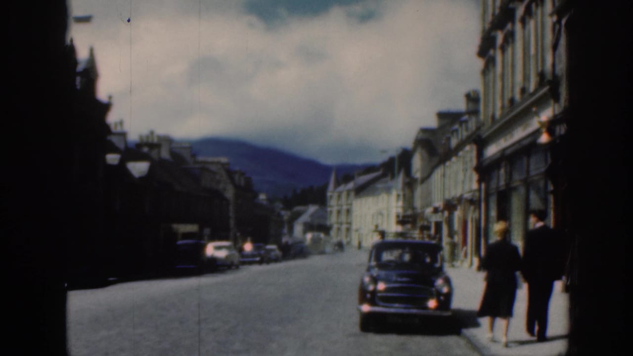 Small-town street with pedestrians, stone buildings, and a dark mid-century car approaching.