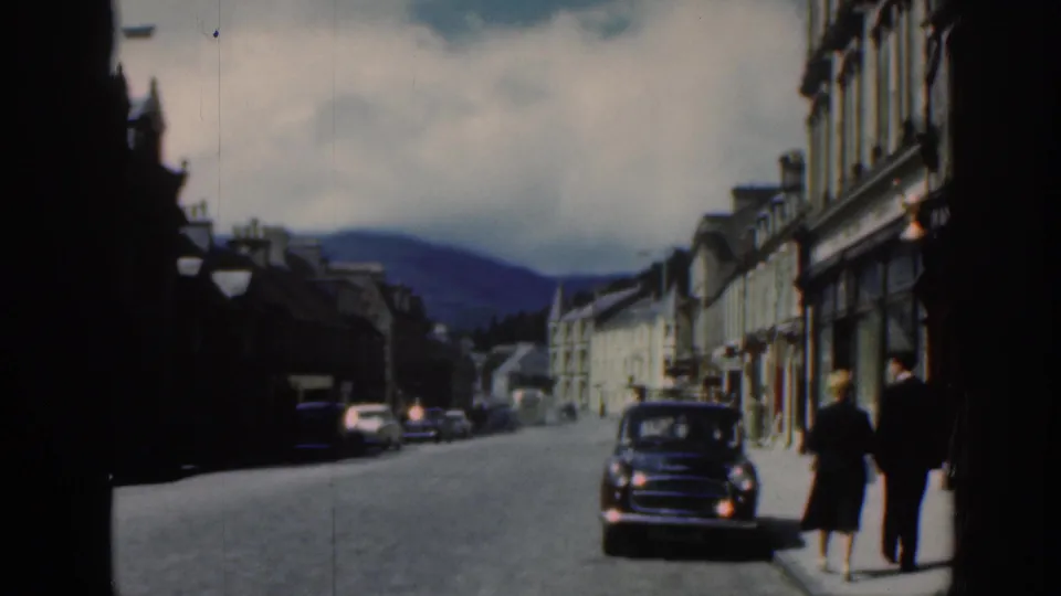 1961 Scotland home-movie frame showing a quiet town street, pedestrians on the road, stone buildings, and a dark car near the curb.