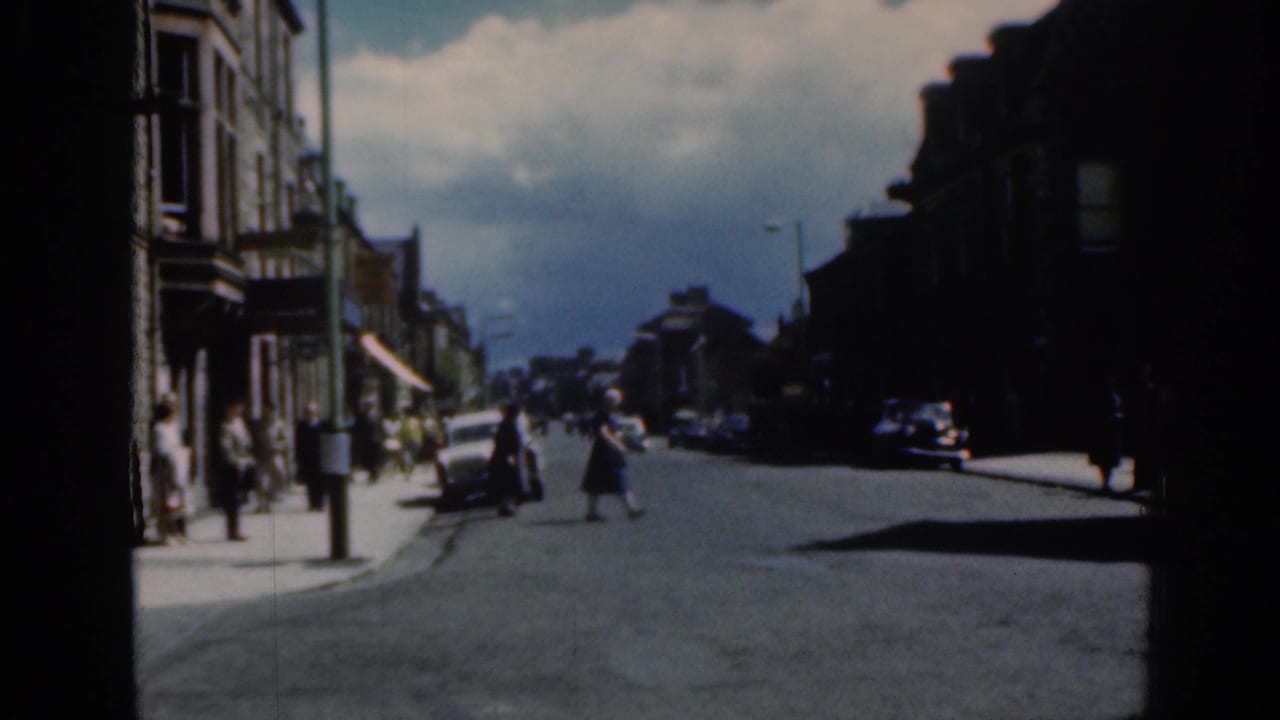 Street corner with people walking, stone buildings, and cars along a broad road.