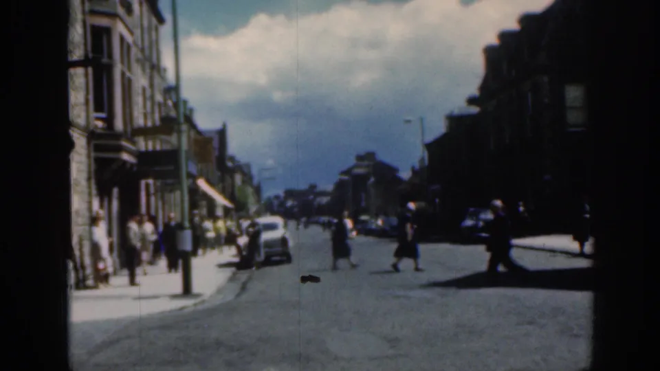 1961 Scotland home-movie view of pedestrians crossing a town street near stone buildings, awnings, and mid-century cars.