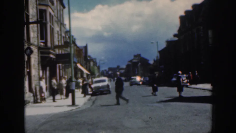 1961 Scotland street scene with pedestrians crossing and walking near a white car, stone buildings, and a cloudy sky.