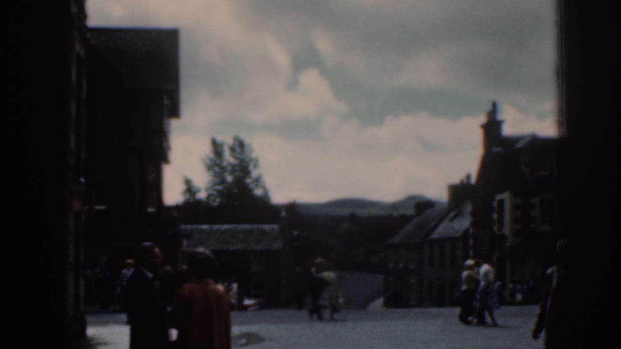Pedestrians in a shadowed town square with buildings and cloudy sky beyond.