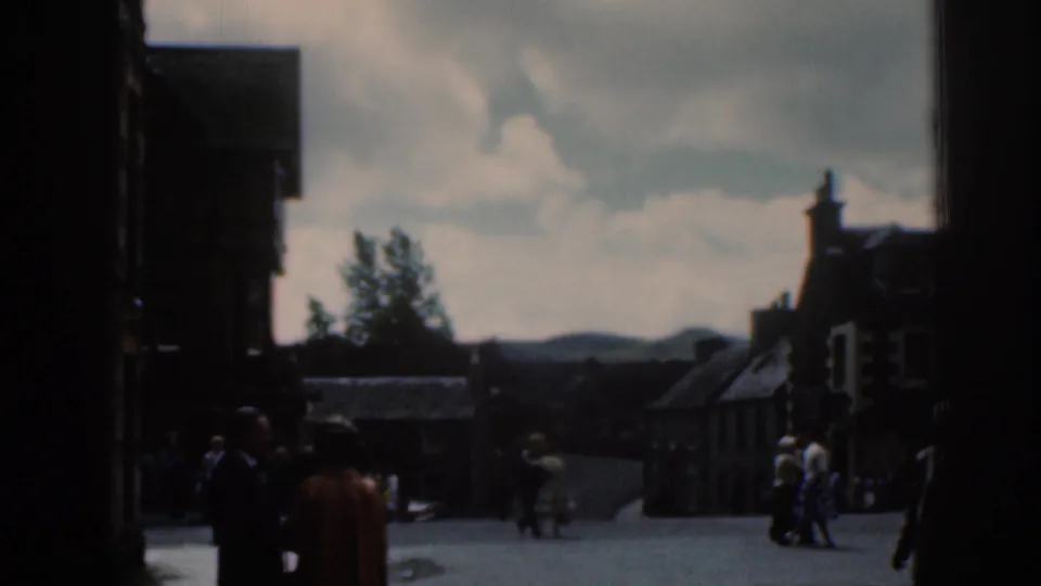 1961 Scotland home-movie frame showing people walking through a town square, partly obscured by dark foreground edges and backlit buildings.