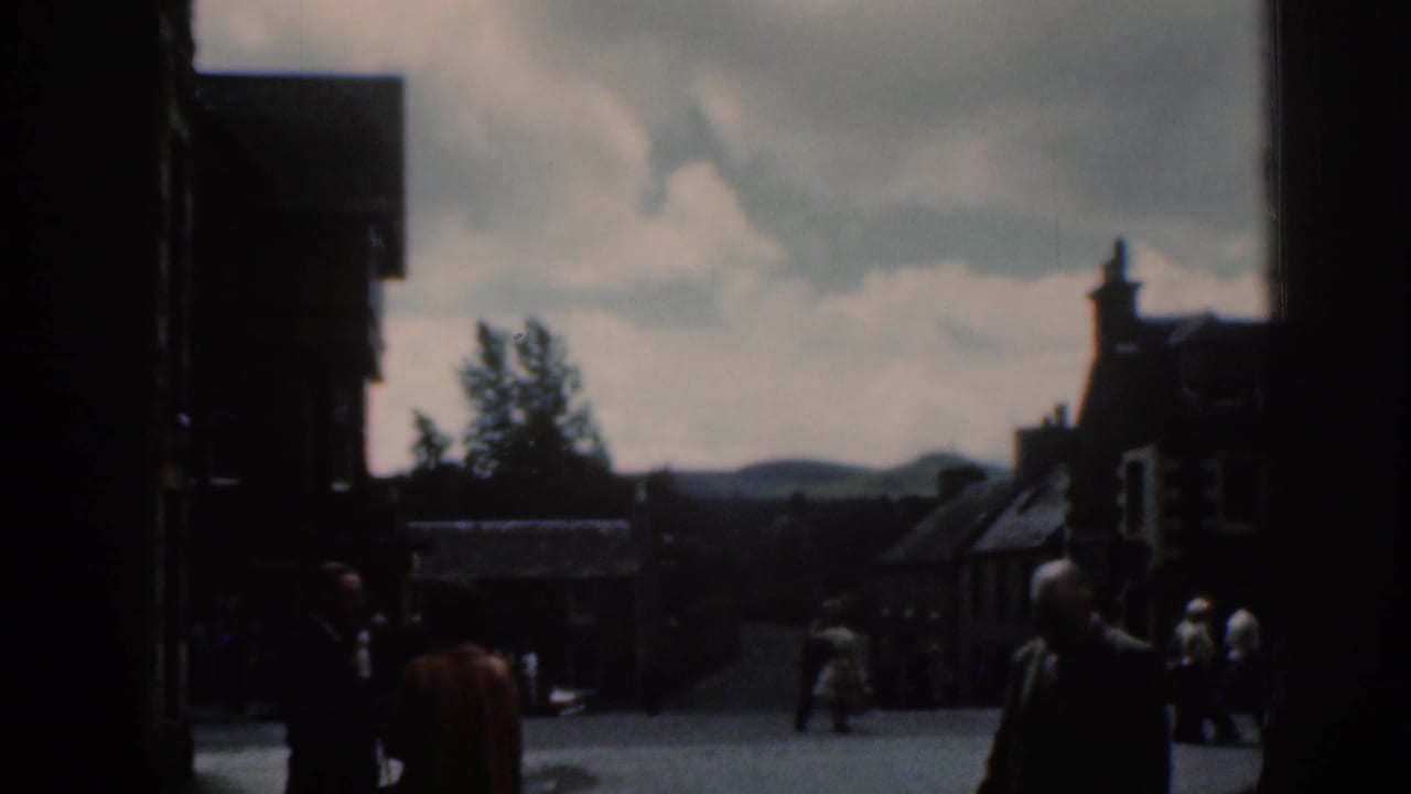 1961 Scotland street scene with adults in period clothing crossing a public square, with buildings, sky, and distant hills in soft focus.