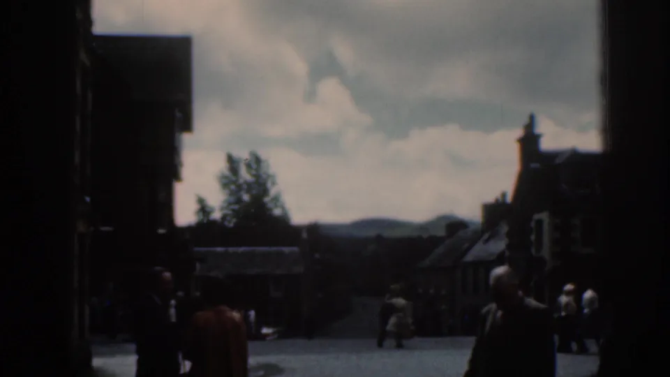 1961 Scotland street scene with adults in period clothing crossing a public square, with buildings, sky, and distant hills in soft focus.