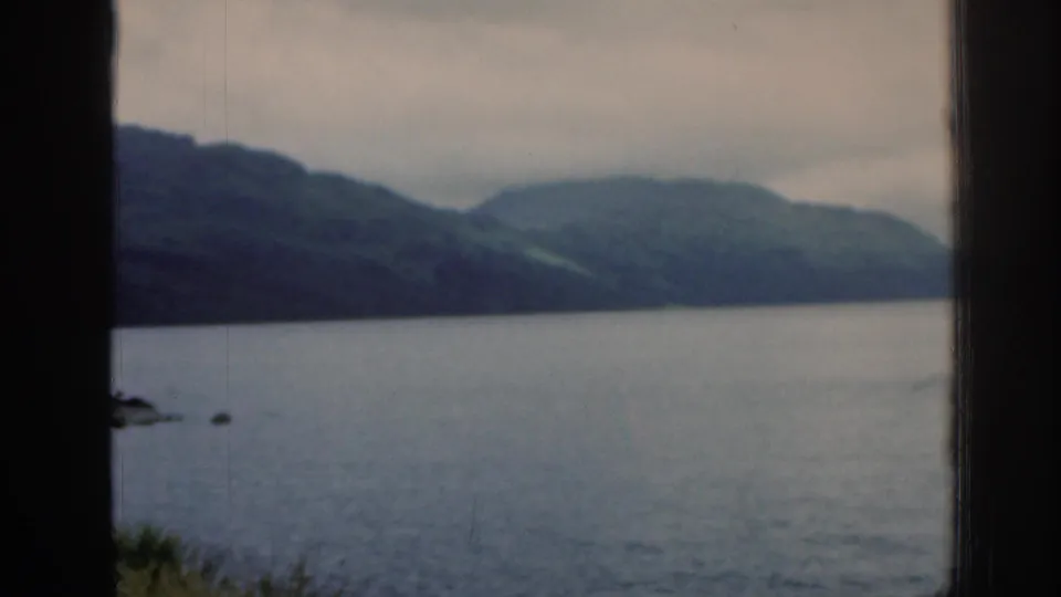 1961 Scotland home-movie view of a broad loch and rolling hills, seen through dark side framing with muted color and soft grain.