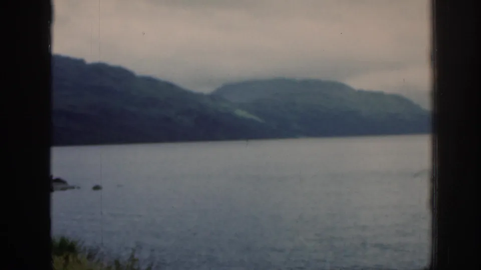 1961 Scotland landscape frame showing calm loch water, green hills, and cloudy sky, held in a visitor-shot composition from a shaded opening.