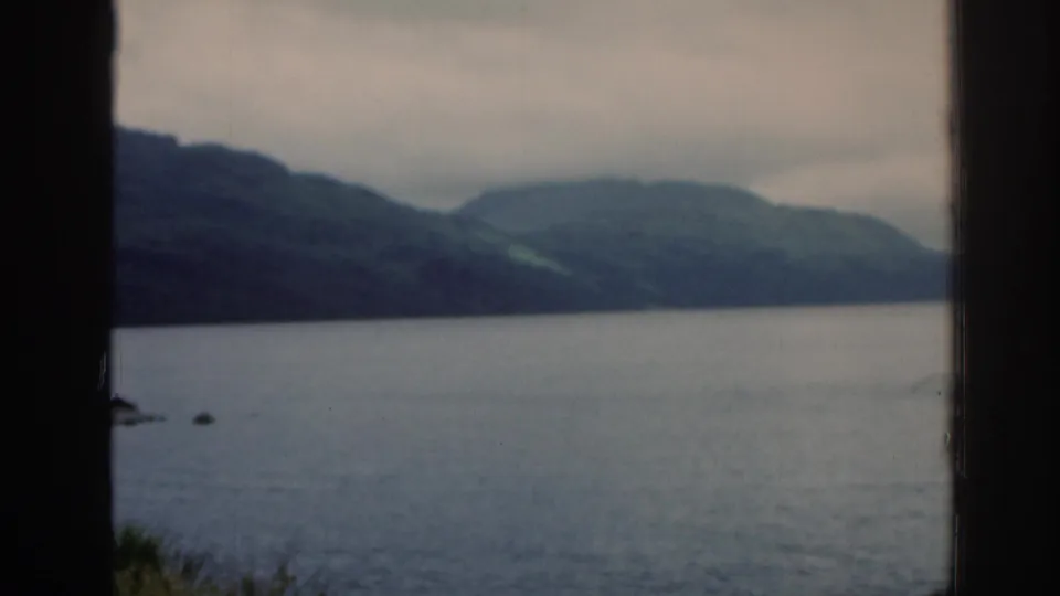 1961 Scotland 8mm frame showing a calm loch, distant green hills, cloudy sky, and dark vertical window framing.