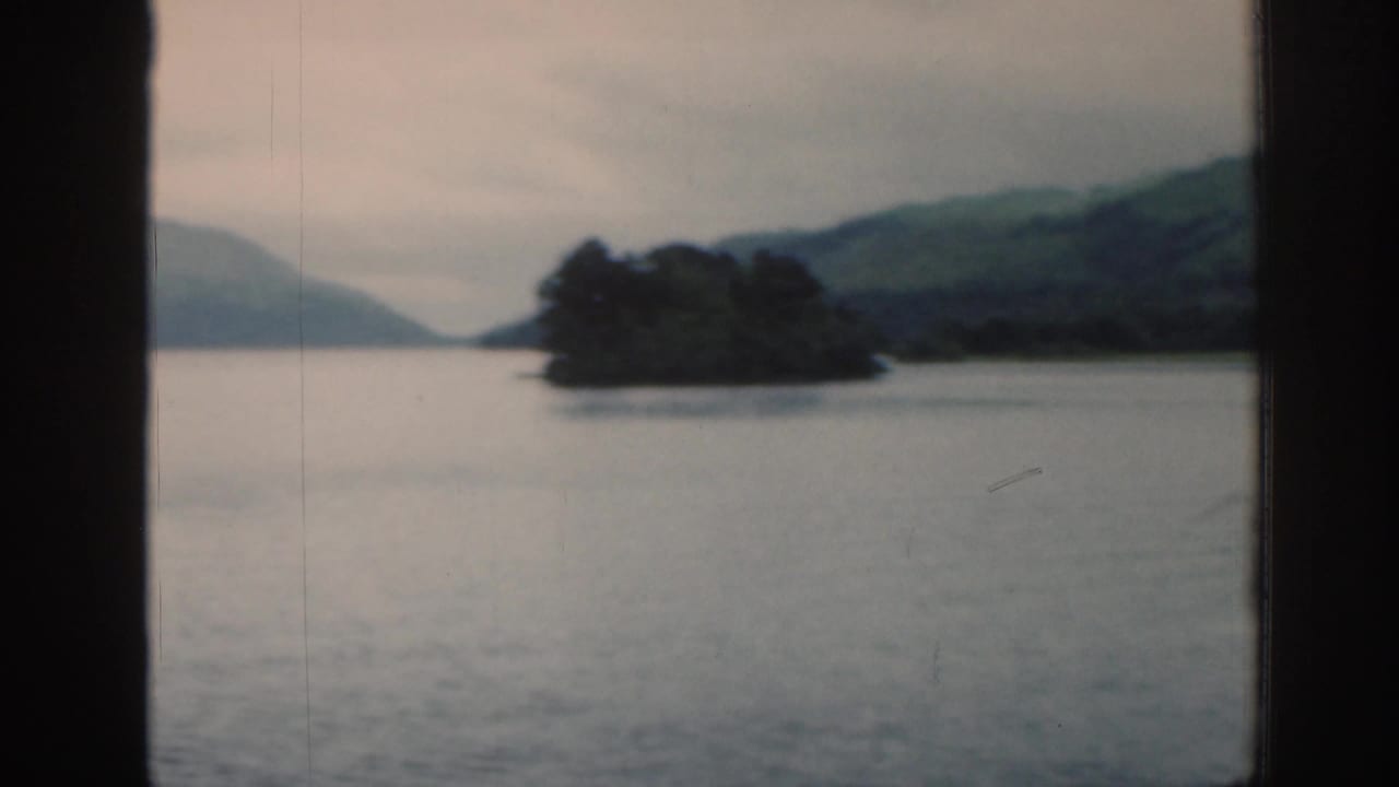 Small wooded island in a loch with hills and cloudy sky behind it.