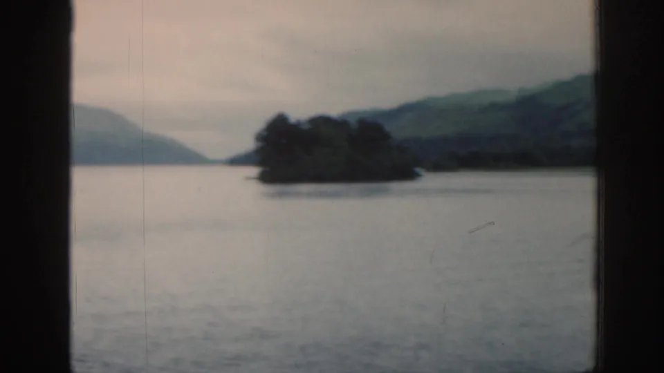 1961 Scotland travel frame with a tree-covered island in a still loch, backed by green hills and an overcast sky.