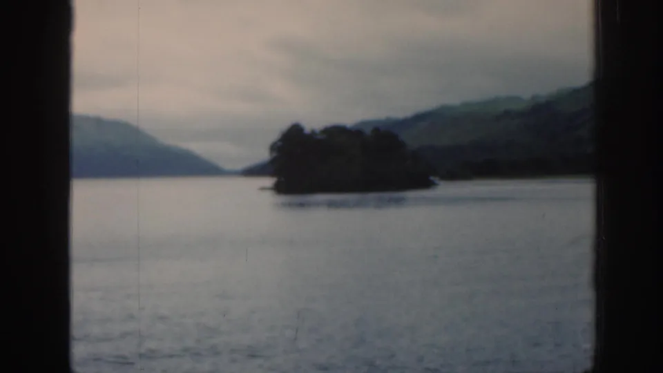 1961 Scotland frame showing a soft-focus loch landscape with a wooded island, distant hills, and grey cloud cover.