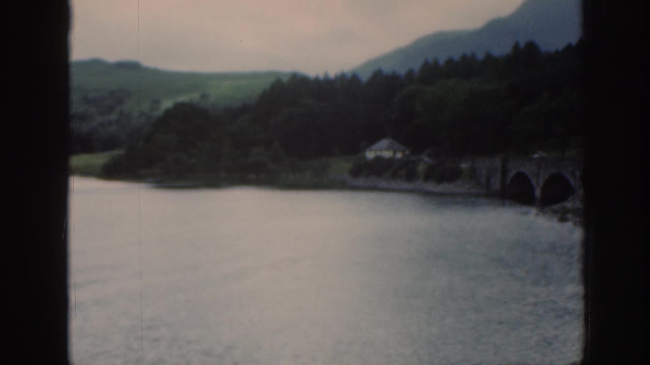 1961 Scotland home-movie frame with a soft lake view through window bars, showing trees, hills, a small building, and stone bridge.