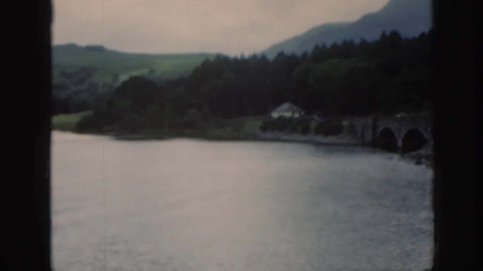 1961 Scotland home-movie frame with a soft lake view through window bars, showing trees, hills, a small building, and stone bridge.
