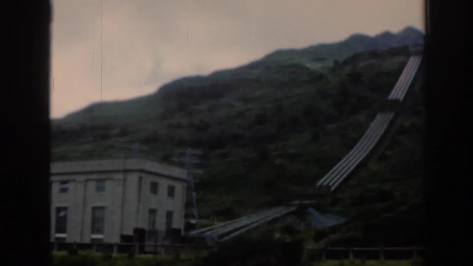 1961 Scotland 8mm frame showing a utility building below a steep green hillside with large penstock pipes rising upward.