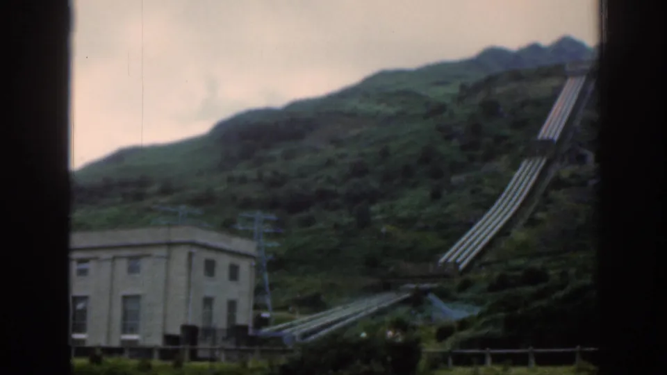 1961 Scotland home-movie frame of a hydroelectric facility area, with a pale building, hillside pipes, green slopes, and distant ridges.
