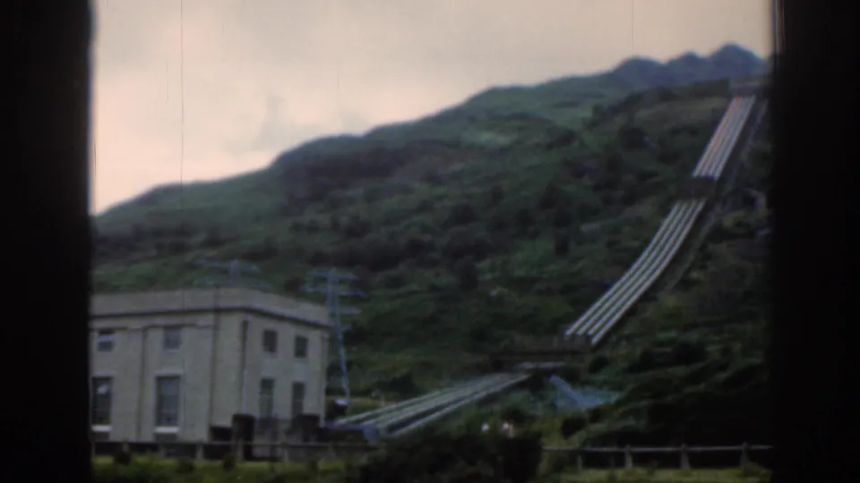 1961 Scotland home-movie frame showing a small power station structure below large hillside pipes and green mountain terrain.