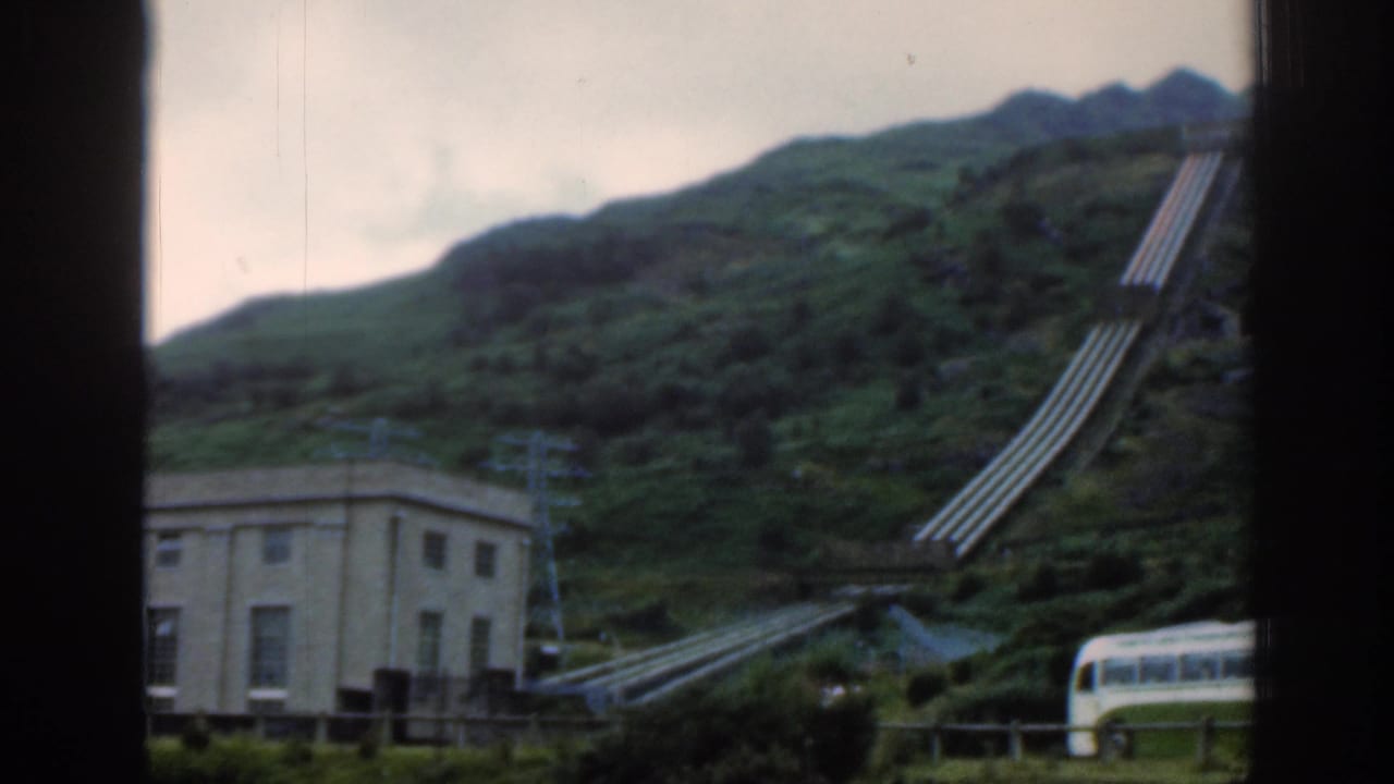 1961 Scotland home-movie view through a window frame, with hillside pipework, a utility building, and a bus partly visible at right.