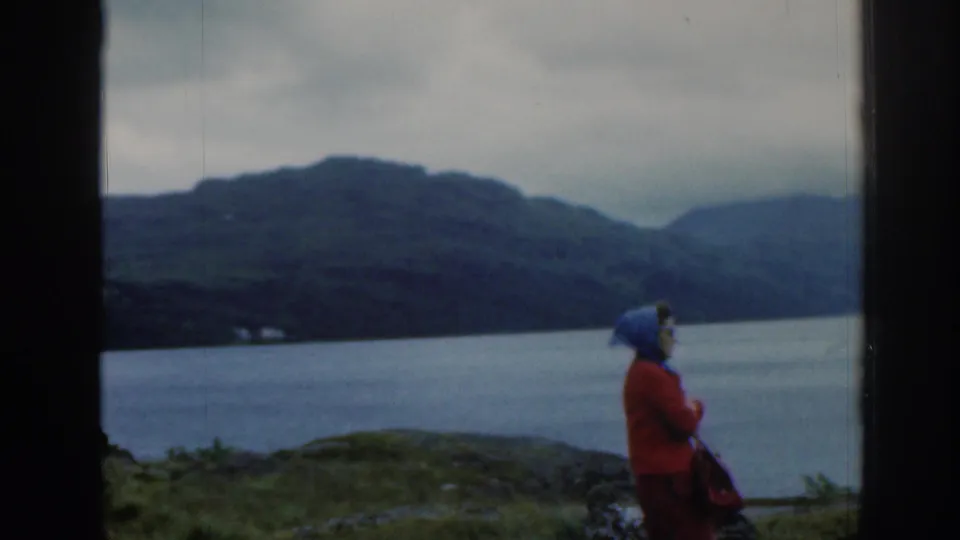 1961 Scotland home-movie frame of a woman in a red coat and blue headscarf standing near a loch with mountains beyond.