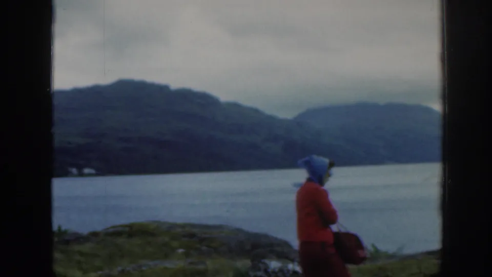1961 Scotland home-movie view of a woman in a red coat beside open water, framed by dark vertical window edges and misty mountains.