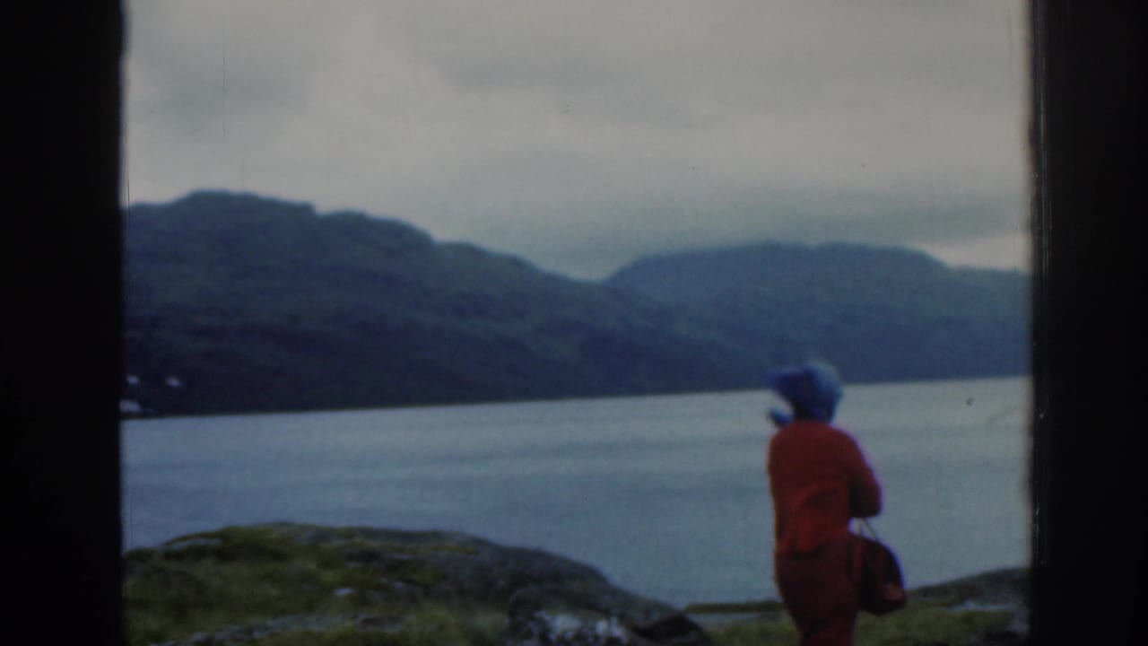1961 Scotland home-movie frame of a woman in a red coat and blue headscarf looking across a lake toward misty hills.