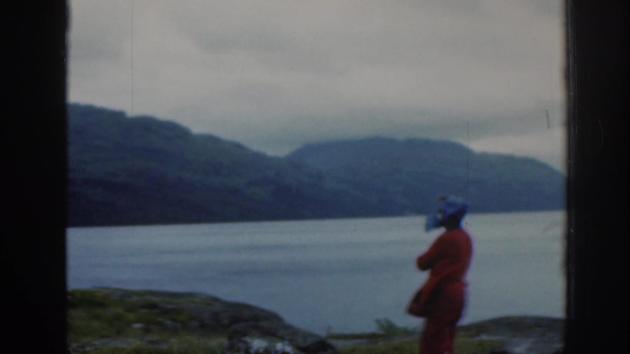 1961 Scotland frame showing the woman in red in profile beside a calm lake, with rolling hills under an overcast sky.