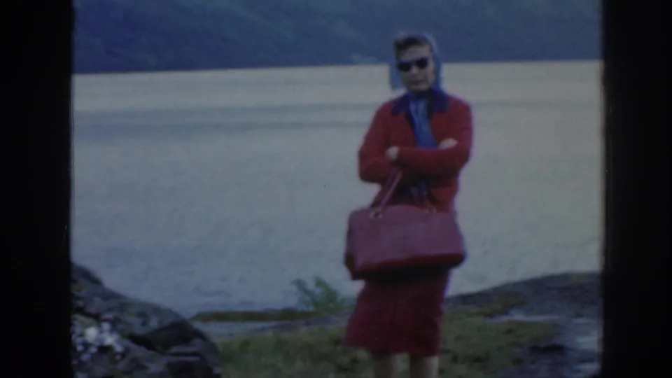 1961 Scotland home-movie frame of the woman facing the camera in sunglasses, red coat and blue scarf, with lake and hills behind.