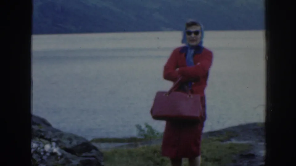 1961 Scotland frame of the woman standing with arms crossed, wearing sunglasses and holding a red handbag beside a lake.