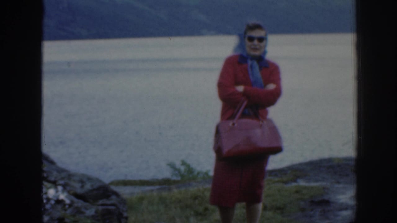 In 1961 Scotland home-movie footage, a woman in red outerwear, sunglasses, and a blue scarf stands near a loch with hills behind her.